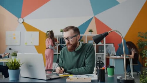 Serious Guy Using Laptop and Writing Notes Working in Open Space Office with Colleagues