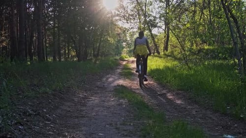 Young Woman on a Bicycle Rides Along Green Forest Path in Sunny Summer Day