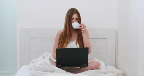 Young Woman Freelancer Is Sitting on the Bed Drinking Coffee and Working on a Laptop, Morning Time