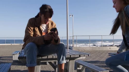 Front view of a Caucasian and a mixed race girl sitting on a table seaside