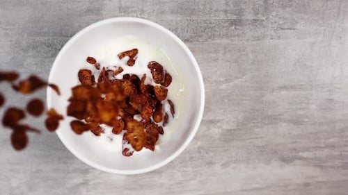 Top View of Pouring Cornflakes with Chocolate Flavor Into a White Bowl with Milk in Slow Motion