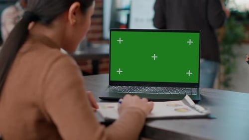 Woman Reviews Charts Beside Laptop with Green Screen