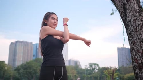 Asian sport woman stand and do arm stretching in green park or garden with cityscape as background