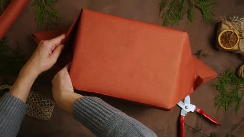 Hands Wrapping Gift on Table With Holiday Decorations