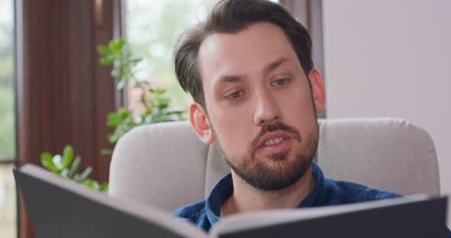 Close Up Portrait of Young Bearded Man Reading Book While Sitting in White Chair Young Man Relaxing