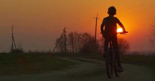 A Boy Is Riding a Bicycle on a Pathway at Sunset. Filming From the Back.