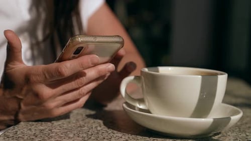Nahaufnahme einer weißen Tasse mit einer Untertasse und einer weiblichen Hand mit einem Telefon