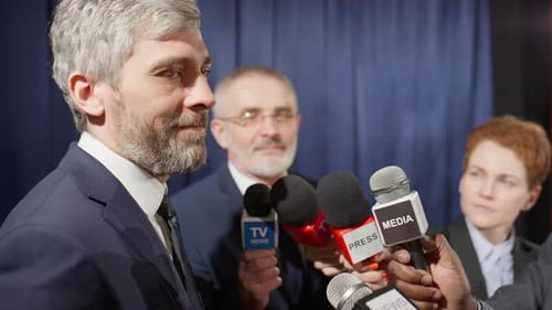 Man Speaking at Press Conference Surrounded by Microphones
