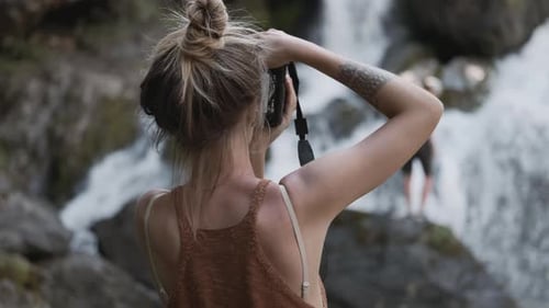 Young Paparazzi Girl Photographs a Waterfall Standing on Stones in the Jungle