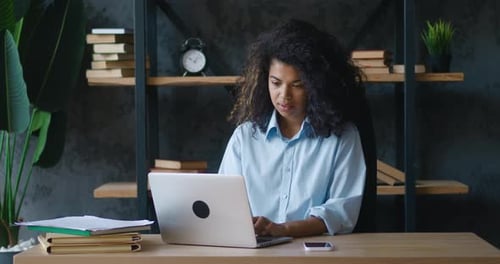 African American Young Woman with Curly Hair Working on Laptop Looking at Smartphone at Office