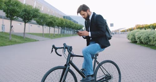 Hombre barbudo sentado en bicicleta durante la parada en el sendero peatonal en el fondo del edificio de la ciudad