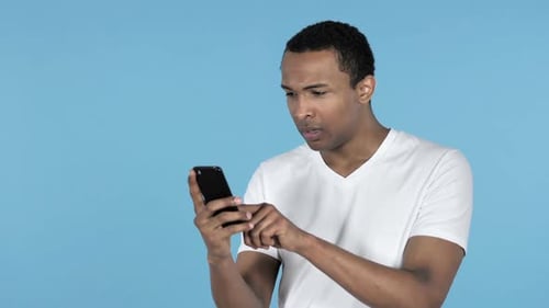 Joven africano que reacciona a la pérdida y usa un teléfono inteligente de fondo azul
