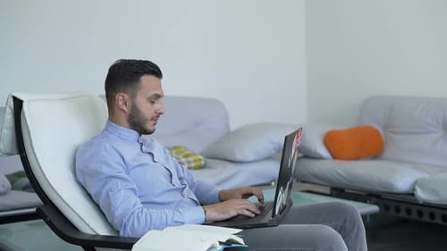 Young Bearded Businessman Sitting in Home Office at Table, Writing Documents and Working on Laptop