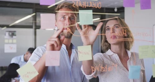 Professional businesswoman and businessman discussing over memo notes on glassboard in modern office