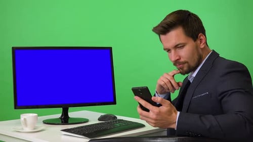 A Young Businessman Sits in Front of a Computer and Works on a Smartphone - Green Screen Studio