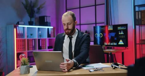 Bearded Businessman Sitting at His Workplace in Evening Office and Working on computer