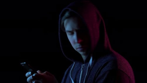 A Young Man in a Hood with a Phone in His Hands. Hacker Makes a Hack Through the Phone. Blue and Red