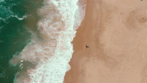 Vista superior de un surfista solitario camina por la playa mientras el mar salvaje sin fin aplastando las olas en la arena