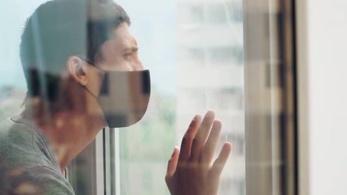 Guy in medical mask fit to closed window during quarantine and isolation at home due to coronavirus