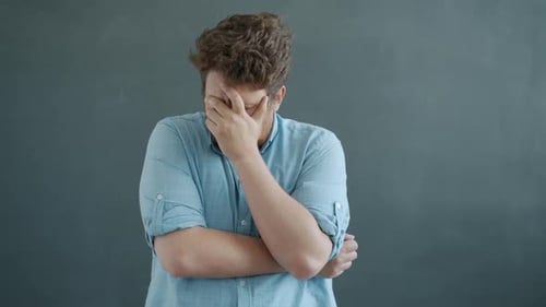 Worried Man Stands Against Grey Background, Hand to Face