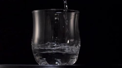Clean water falling into a transparent glass, over a black background. Closeup slow motion shot.