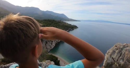 Boy Traveller with Arm Raised on Top of Mountain Looking at View of Adriatic Sea and Rocky Mountains