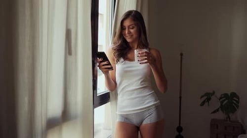 Smiling Woman Uses Smartphone at Home Near Window