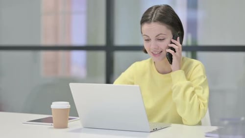 Young Woman Talking on Smartphone While Using Laptop in Office