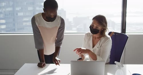 Two diverse female colleagues wearing face masks looking at laptop and discussing in office