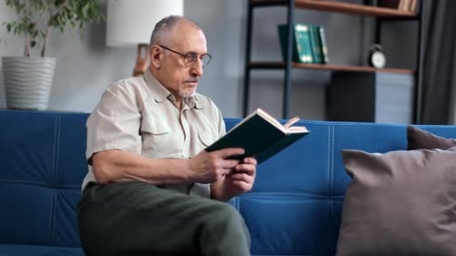 Enthusiastic Aged Man Reading Interesting Book Turning Page Sitting on Blue Couch Time for Yourself