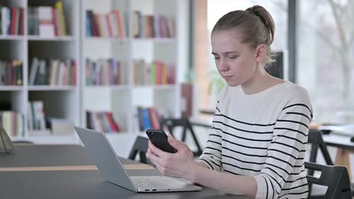 Smartphone Use By Young Woman with Laptop in Library