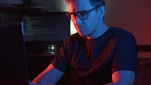 Man Types at Laptop in Dark, Red and Blue Lit Room