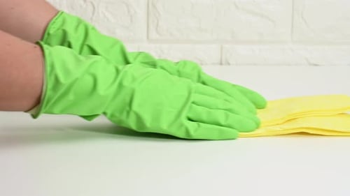 hand in a green rubber glove holds a yellow rag and wipes a white table