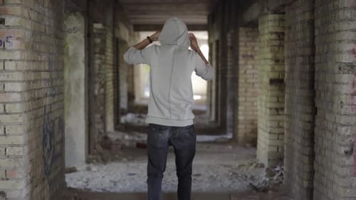 Back View of Young Male Bandit Walking Along Abandoned Ruins and Putting on Hood. Wide Shot of