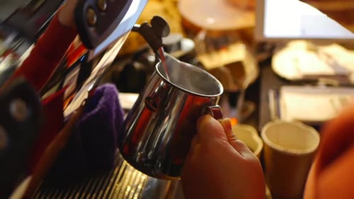 Close up on hands of coffee barista using coffee machine to add hot milk into silver jug