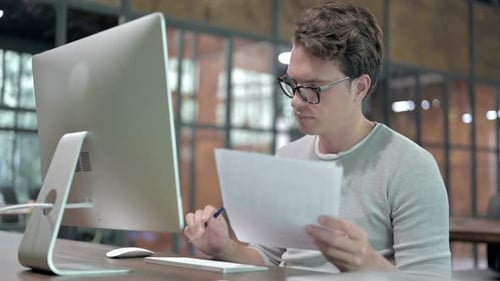 Young Adult Reviews Document at Computer in Office