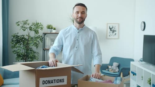 Young Man Collecting Things for Donation Putting Clothes in Cardboard Box Indoors at Home