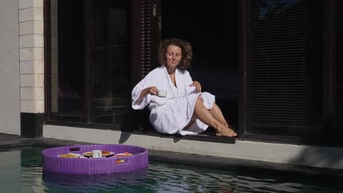 Young Caucasian Woman in a Robe Sitting on the Edge of Swimming Pool Taking a Cup of Coffee From the