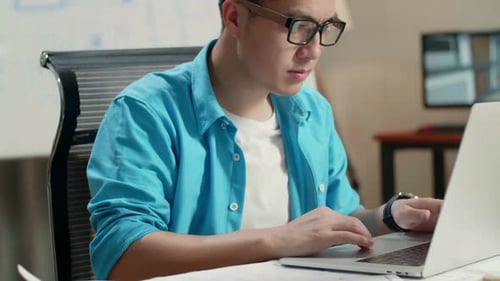Close Up Of Asian Man Engineer Looking At Blueprint While Typing On A Laptop At The Office
