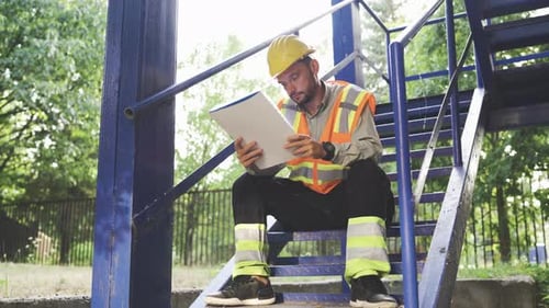 Tired Civil Engineer in Helmet and Safety Vest Sitting on Stairs with Blueprints