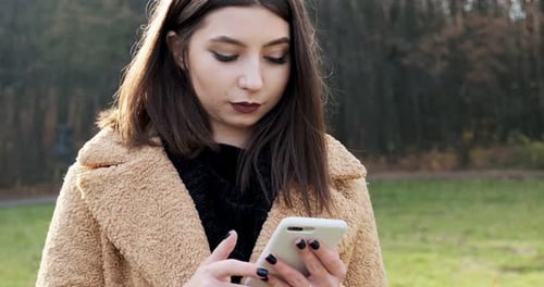Portrait of Young Attractive Woman Smiling and Looking at Smartphone on the Green Lawn at Autumn