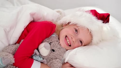 Menina sorridente de chapéu de Papai Noel e pijama de Natal deitada na cama com ursinho de pelúcia em casa
