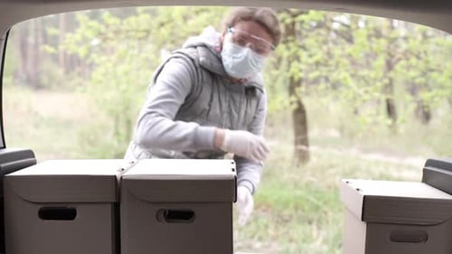 A Volunteer in a Protective Suit Loads Boxes Into the Trunk of a Car