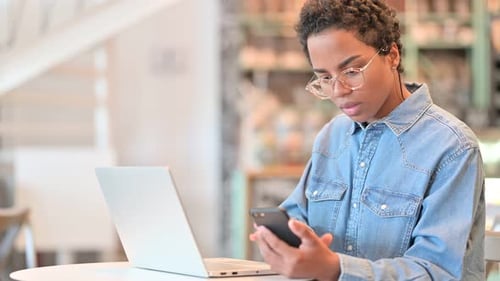 Hardworking African Woman Using Smartphone and Laptop at Cafe