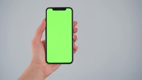 Female Hands Holding a Smartphone with a Green Screen on a Gray Background. Chroma Key. Copy Space