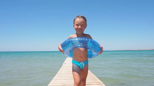 Little Boy in Swimming Trunks Runs Along a Wooden Pier for the Camera Against the Background of the