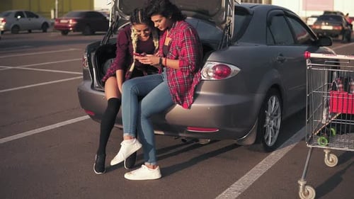 Two Women Using Phone Sitting on Car Trunk