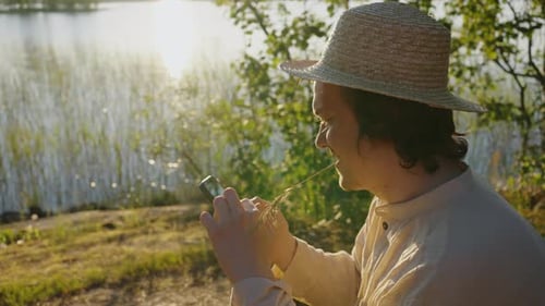 Young Man Surfs Internet on Smartphone Sitting on River Bank