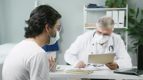 Elderly Grayhaired Doctor Speaking with Young Patient About Treatment in Hospital Wearing Protective