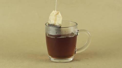 Schwarzer Teebeutel zum Heben und Fallenlassen in einen transparenten Glasbecher zum Aufbrühen von Tee isoliert auf Beige